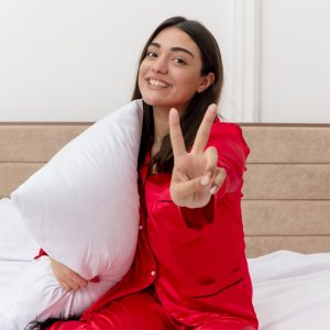 young beautiful woman in red pajamas sitting on bed with pillow looking at camera smiling showing v-sign in bedroom interior on light background